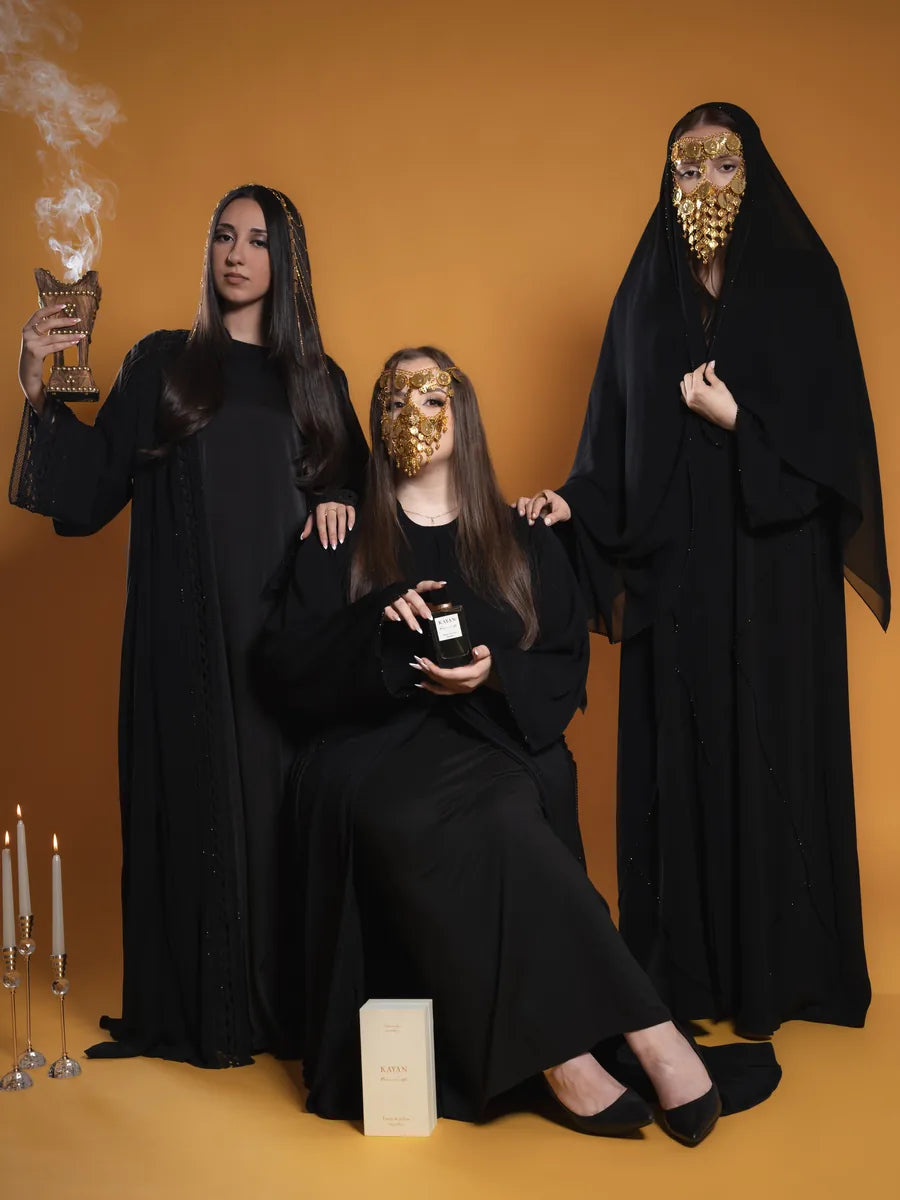 Three women in black abayas, two wearing a traditional gold face mask; one holds a smoking incense burner. Maison KAYAN bottle and box in the foreground.