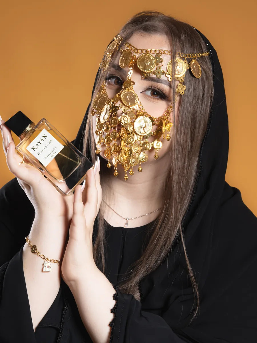 A woman in a black abaya wearing a traditional gold face mask, holding the Maison KAYAN Premier Souffle bottle.